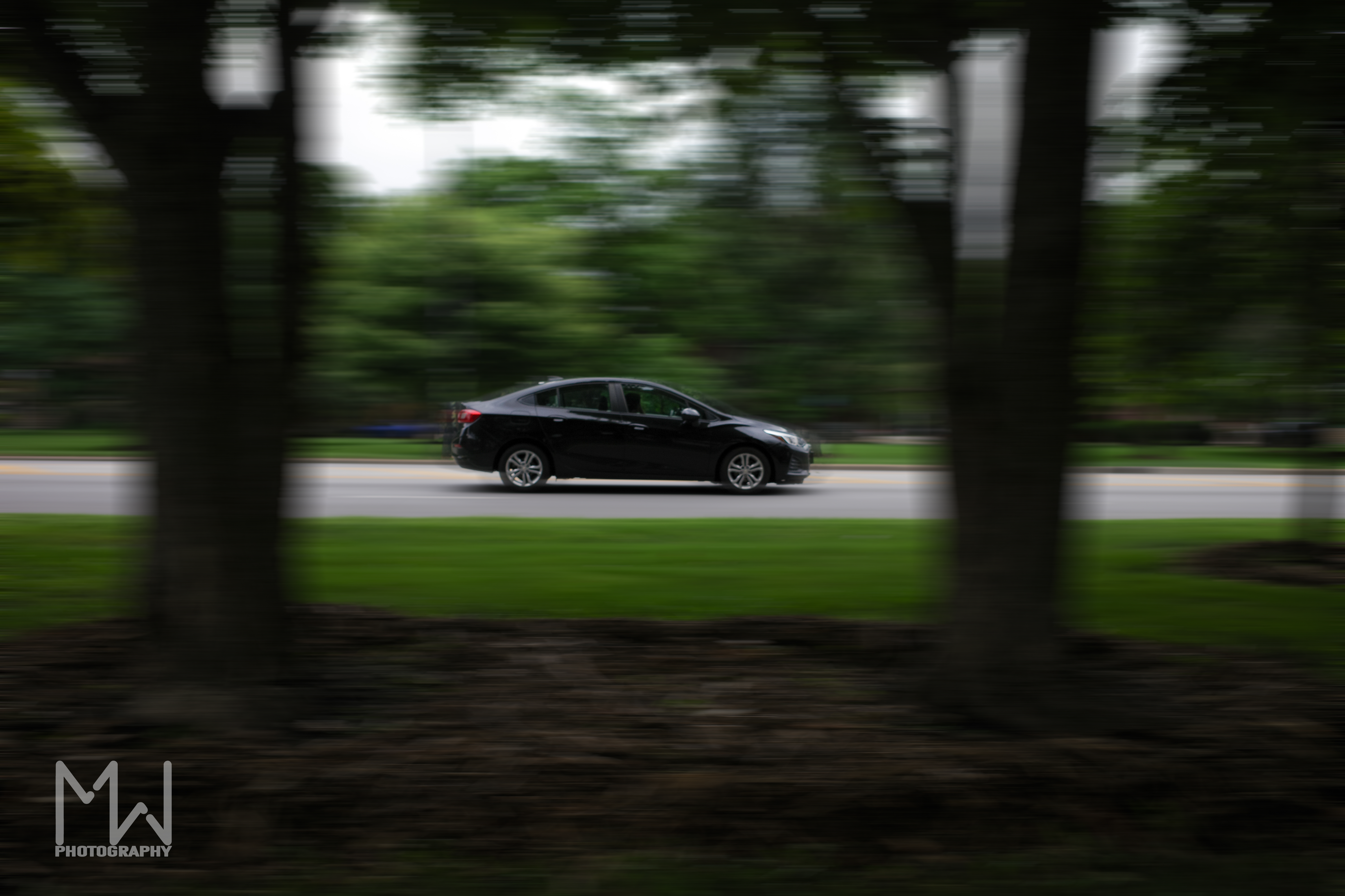 A photo of a car driving in between two trees in the city with greenery in the background and motion blur