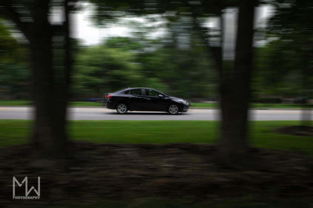 A photo of a car driving in between two trees in the city with greenery in the background and motion blur